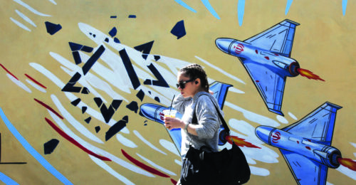 Dziewczyna idąca z prawej do lewej strony kadru, ubrana w jasnoszarą bluzę, z dużą czarną torbą przewieszoną przez ramię. Osoba trzyma w dłoni plastikowy kubek z napojem i słomką, ma włosy zaplecione w warkocz oraz nosi ciemne okulary przeciwsłoneczne. W tle znajduje się duży, kolorowy mural przedstawiający stylizowane samoloty w odcieniach niebieskiego i szarości, namalowane na żółtym tle