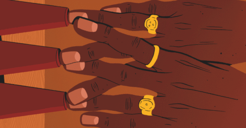 Ilustracja przedstawia zbliżenie dwóch dłoni ułożonych na stole. Palce spoczywają na trzech odwróconych, czerwonych stożkach ustawionych w rzędzie. Na palcach widoczne są złote pierścionki o prostych, okrągłych kształtach. Skóra dłoni ma ciemny odcień, a paznokcie są krótko obcięte. Tło i powierzchnia stołu utrzymane są w ciepłych, brązowych tonach