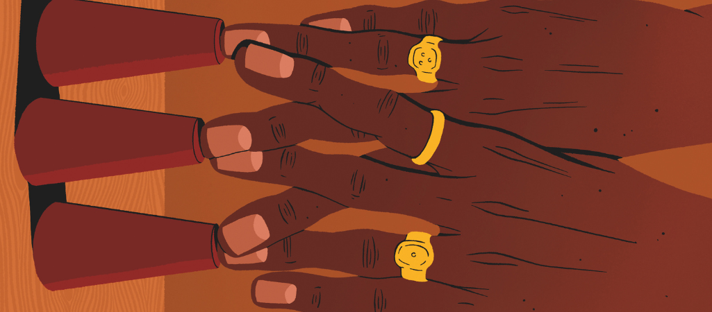 Ilustracja przedstawia zbliżenie dwóch dłoni ułożonych na stole. Palce spoczywają na trzech odwróconych, czerwonych stożkach ustawionych w rzędzie. Na palcach widoczne są złote pierścionki o prostych, okrągłych kształtach. Skóra dłoni ma ciemny odcień, a paznokcie są krótko obcięte. Tło i powierzchnia stołu utrzymane są w ciepłych, brązowych tonach