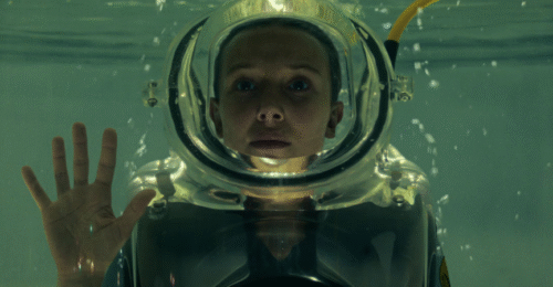 Kadr z serialu Stranger Things. Dziewczynka z krótkimi włosami ubraną w przezroczysty strój nurka jest zanurzona w zbiorniku z wodą. Jedną rękę ma uniesioną